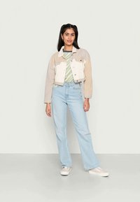 Casaco de ganga bege e branco, cortado, sobre uma camisa riscada verde, combinado com umas calças de ganga azuis claras de perna reta e sapatilhas brancas.