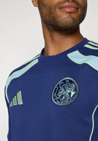 Mørkeblå treningsskjorte med lyseblå detaljer. Har rund hals, brodert Ajax-logo, og tre grønne striper på skulderen.