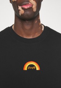 Gros plan d'une personne atteinte de vitiligo sur les lèvres et le cou, portant une chemise noire avec un petit arc-en-ciel brodé jaune, orange et rouge et le texte "Wrangler".