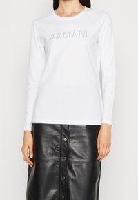 Valkoinen pitkähihainen paita, johon on painettu "ARMANI"-logo strasseilla etupuolelle, yhdistettyinä korkea vyötärö mustaan nahkahameeseen, jossa on napit.