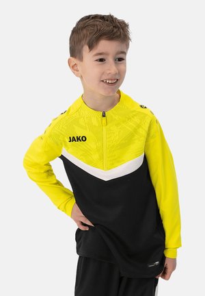 Geel en zwart sporttop met een ritskraag, voorzien van een geometrisch patroon en een witte chevronaccent over de borst. Merklogo aan de voorkant.