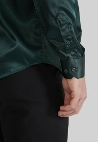 Chemise satinée vert foncé à manches longues, dotée de poignets à boutons et d'une texture lisse et brillante. L'ourlet est légèrement courbé au bas.