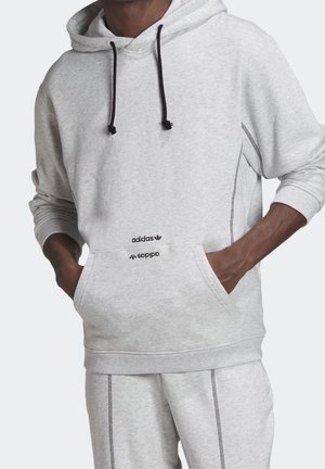 Persoon draagt een lichtgrijze Adidas-hoodie en bijpassende joggingbroek met zwarte trekkoorden en zichtbare naden, handen in de voorzak van de hoodie.