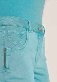 Turquoise skinny jeans met een gladde textuur, voorzien van een klein achterzakje en een zijzak met rits, afgewerkt met contrasterende stiksels.
