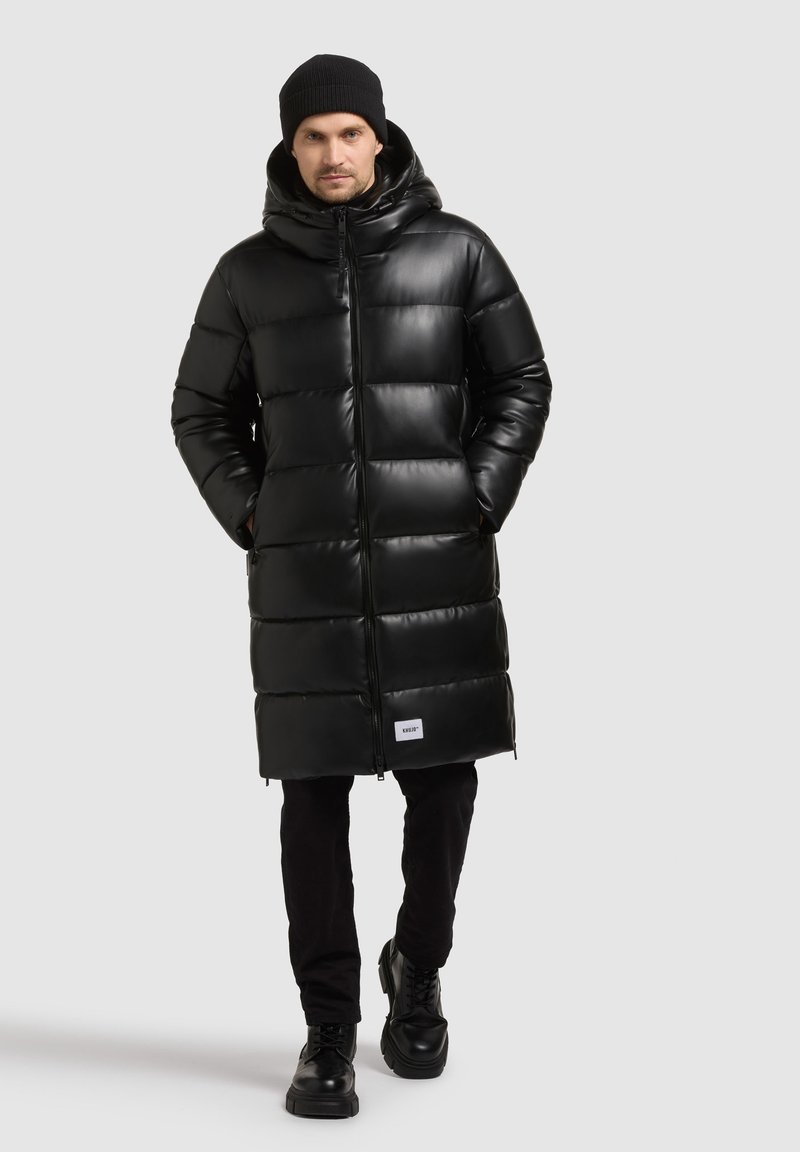 Cappotto imbottito nero con finitura lucida, cappuccio e zip frontale. Presenta sezioni trapuntate verticali e una patch con logo all'orlo.