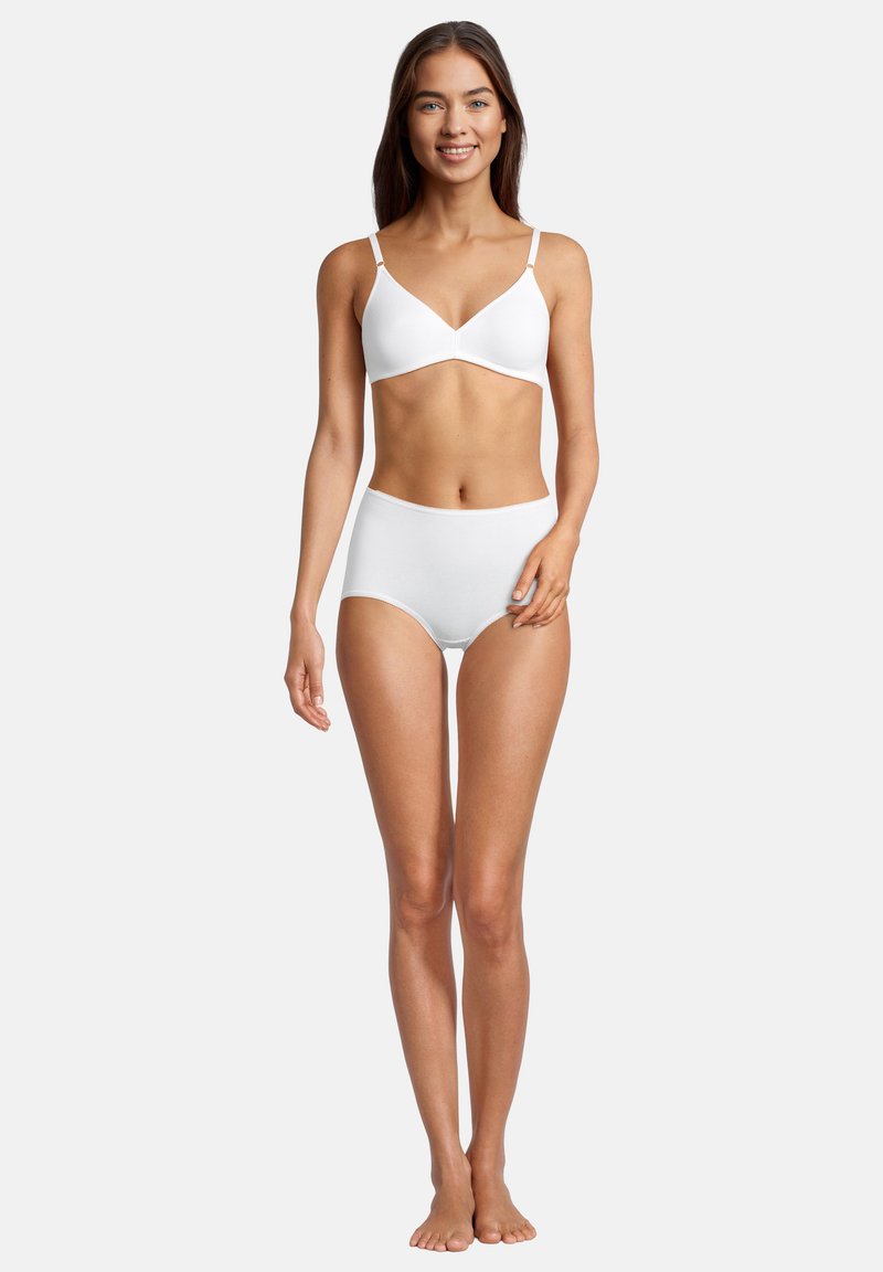 Young woman standing barefoot wearing white cotton bra and high-waisted briefs against plain light background, smiling forward.