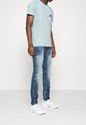 Mann trägt ein hellblaues Lee-T-Shirt, schlank geschnittene, ausgewaschene blaue Jeans und weiße Turnschuhe, steht vor einem schlichten weißen Hintergrund.