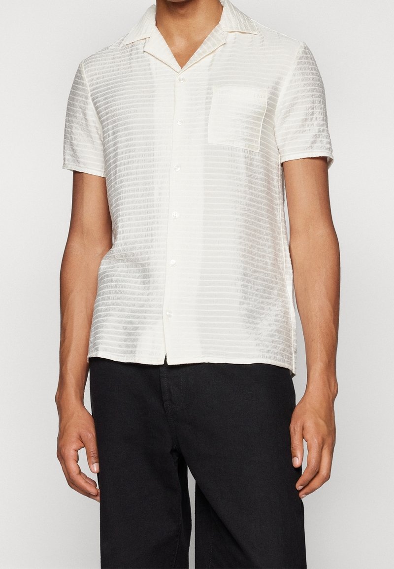 Homme portant une chemise boutonnée blanche texturée à manches courtes avec une poche poitrine et un pantalon noir sur un fond uni.