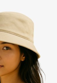 Cappello a secchiello beige in tessuto strutturato, con tesa larga e design strutturato. Finitura liscia, mostrato parzialmente indossato.