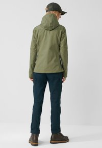 Olivgrön vattentät jacka med huva, kombinerad med mörkblå byxor och bruna kängor, som visar en elegant design och praktisk passform.
