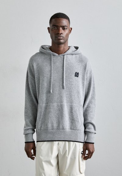 Grå hoodie lavet af blødt stof, med en frontlomme og ribbet kant. Bemærkelsesværdigt sort logo på brystet og justerbar snoretræk i hætten.
