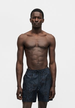 Muž bez trička s tmavou pokožkou a krátkymi vlasmi stojaci pred jednoduchým pozadím, nosiaci tmavé vzorované plavky s šnúrkou.