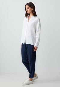 Chemise en lin blanche à boutons avec une coupe décontractée, associée à un pantalon bleu marine ample et des baskets blanches. Design simple, sans motifs supplémentaires.