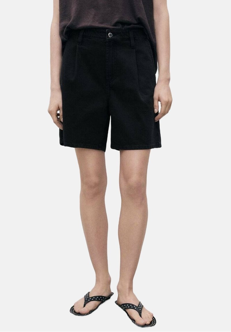 Shorts de mezclilla negra con un ajuste relajado, pliegues delanteros y cierre de botón. Combinados con una parte superior oscura y sandalias negras.