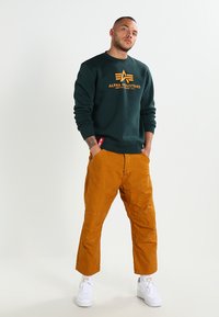Zöld pulóver sárga "Alpha Industries" logóval, narancssárga térdnadrággal, amely textúrált anyagból készült és tapered szabású, fehér edzőcipővel párosítva.