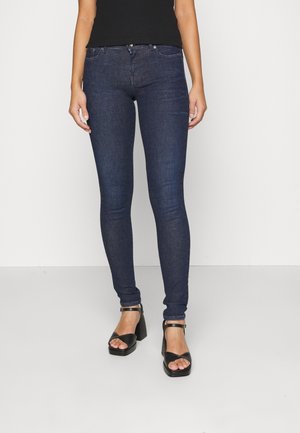 Woman wearing dark blue skinny jeans and black open-toe high-heeled sandals, standing against a plain white background.