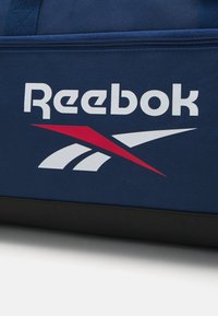 Bolsa de viagem Reebok azul-marinho com um logótipo branco e detalhes em vermelho, apresenta um tecido texturizado e uma base preta. Fecho com zíper na parte superior.