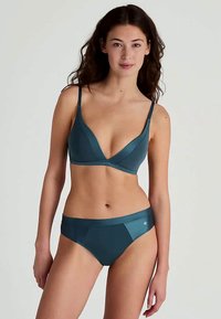 Woman wearing teal-colored bra and matching briefs, standing against a plain light background, facing forward with relaxed expression.