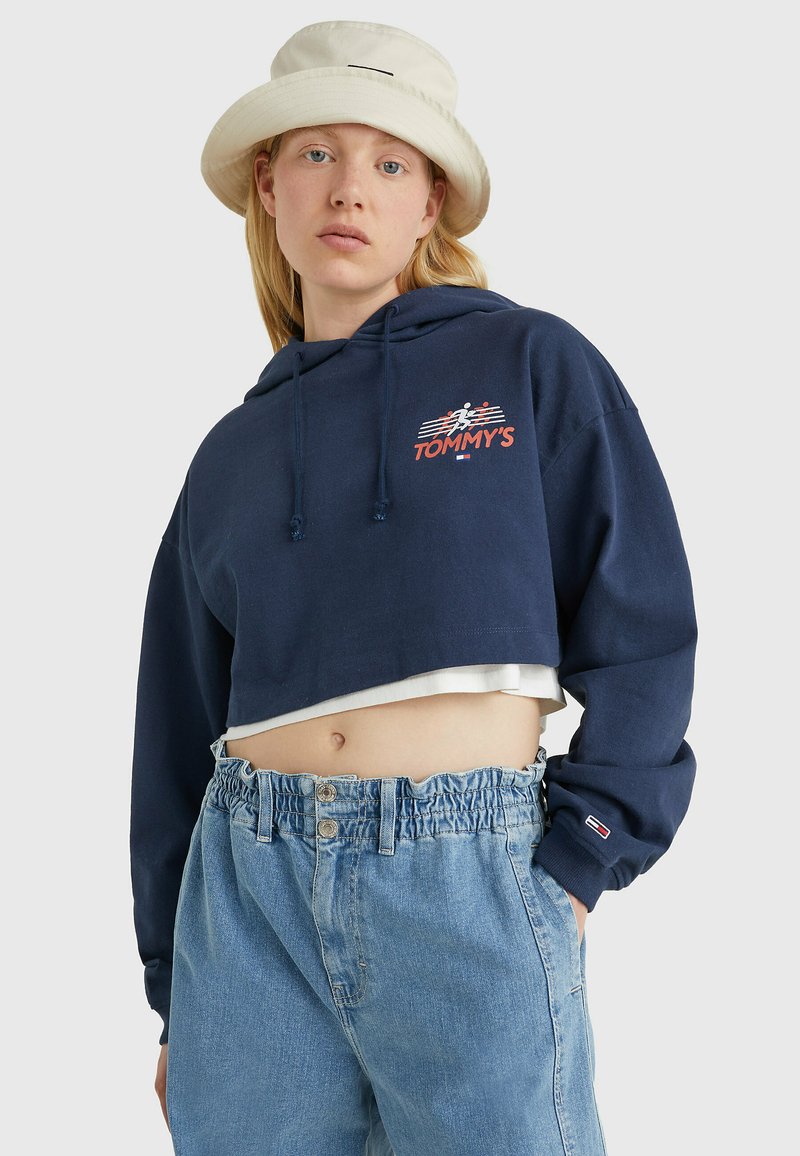 Námornícky skrátený hoody s šnúrkami, s červenobielou grafikou loga "TOMMY'S". V kombinácii so svetlos modrými džínsovými šortkami s vysokým pásom.