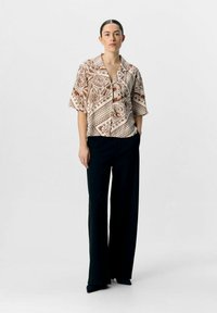 Camicia a maniche corte color beige e marrone con un design a fantasia. Abbinata a pantaloni neri a vita alta. Tessuto liscio, vestibilità comoda.