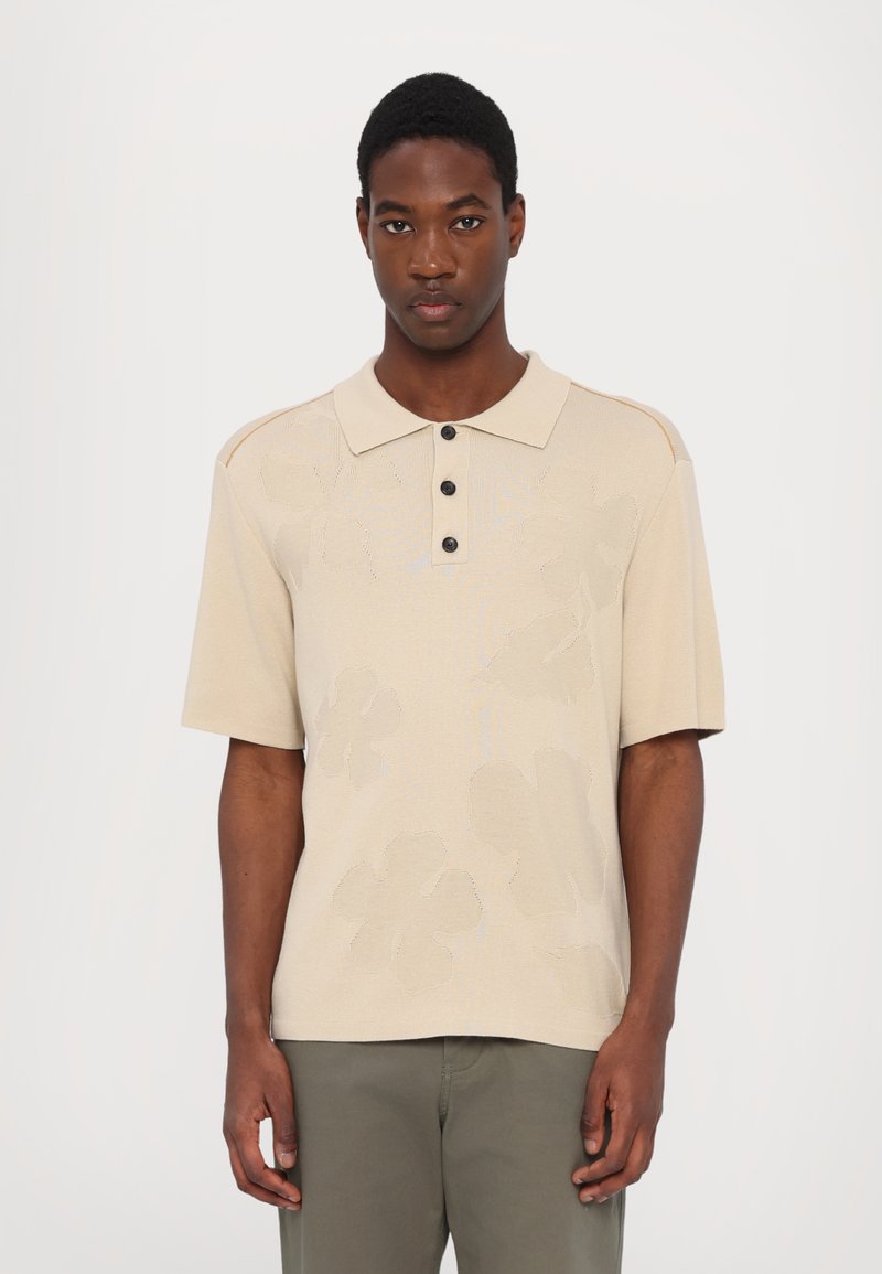Beige stickad polo tröja med korta ärmar, tre svarta knappar och ett subtilt blommönster. I kombination med olivgröna byxor.