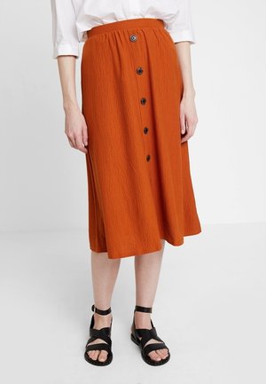 Femme portant une jupe midi orange avec des boutons sur le devant, une chemise blanche retroussée et des sandales plates noires à lanières, debout sur un fond uni.