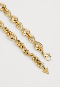 Gouden kettinghalsband met onderling verbonden schakels, een gestructureerd oppervlak en een driehoekige hanger met "GUESS" gegraveerd. Sluiting met een kreeftsklauwsysteem.