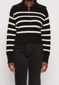 Person, der einen schwarz-weißen, horizontal gestreiften Strickpullover mit halb Zipper und schwarze Hosen trägt, steht vor einem neutralen Hintergrund.
