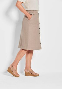Beige, knielanger Rock aus Leinen mit einem taillierten Bund, seitlichen Taschen und Knopfdetails. Kombiniert mit beigen Keilsandalen.