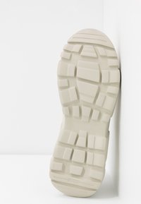 Semelle de botte en caoutchouc beige avec des motifs de bande de roulement profonds et irréguliers pour la traction, photographiée sur fond blanc.