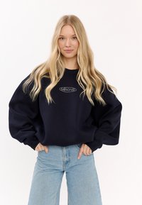 Granatowy, oversize sweatshirt z prążkowanymi mankietami i okrągłym dekoltem, z srebrnym logo na piersi. Połączony z jasnoniebieskimi dżinsami.