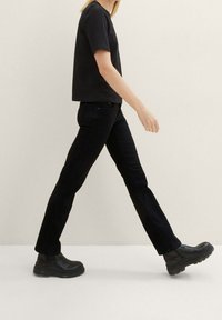 Jeans de mezclilla negros con un corte recto, combinados con una camisa negra de manga corta y botas negras gruesas. Fondo neutro.