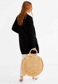 Sac en paille tressé rond avec un motif en spirale, doté de deux poignées circulaires, porté par une personne portant un pull en maille noir et un pantalon clair.