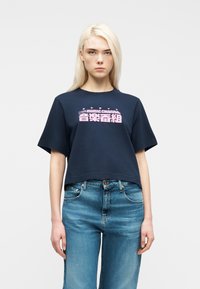 Γυναίκα με μακριά ξανθά μαλλιά που φοράει σκούρο μπλε κοντό μπλουζάκι με ροζ γράμματα και μπλε τζιν, στέκεται μπροστά σε απλό φωτεινό φόντο.