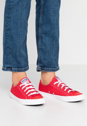 Osoba nosząca czerwone niskie trampki Converse All Star w połączeniu z niebieskimi dżinsami na jednolitym tle.