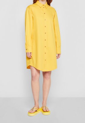 Kvinde iført en gul skjortekjole med lange ærmer og beige plateau-sandaler med gule såler, stående mod en ensfarvet grå baggrund.