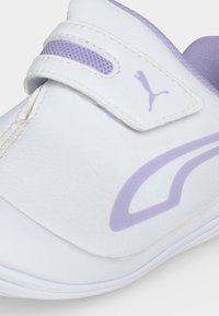 Λευκό και ανοιχτό μωβ αθλητικό παπούτσι Puma με λουράκι βέλκρο και λεπτομέρεια λογότυπου στο πλευρό και στο λουράκι, κοντινή λήψη από μπροστά και πλαγίως.