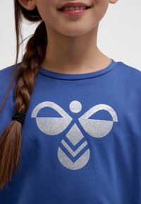 Blaues T-Shirt mit einem großen silbernen Glitzer-Graphic-Design aus geometrischen Formen und Mustern in der Mitte. Weicher Stoff.