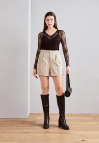 Černý krajkový top s dlouhým rukávem, béžové šortky s vysokým pasem, vysoké černé boty na podpatku po kolena a černá kulatá kabelka na dřevěné podlaze.