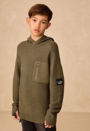 Jeune garçon aux cheveux bruns courts portant un sweat à capuche en maille vert olive avec une poche zippée sur la poitrine et un patch sur la manche.