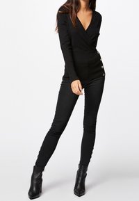 Kobieta w dopasowanej, czarnej bluzce z długim rękawem, z wysokim stanem czarnych spodni z guzikami po bokach oraz czarnych botkach za kostkę.