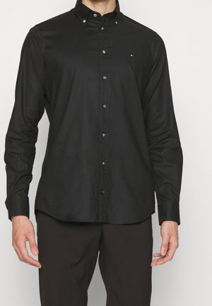 Uomo che indossa una camicia nera a maniche lunghe con bottoni e un piccolo logo ricamato sul petto sinistro e pantaloni scuri.