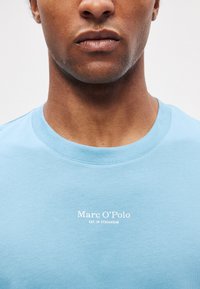 Helehall sinine puuvillane T-särk, millel on ümargune kaelus ja valge trükitud logo "Marc O'Polo EST. IN STOCKHOLM" rinnal. Sile tekstuur.