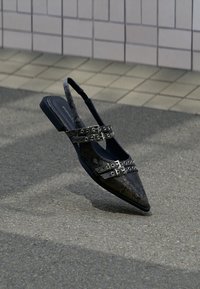 Γυναικείο παπούτσι slingback με μυτερή μύτη σε μαύρο τεχνητό δέρμα φιδιού, με μεταλλικές αγκράφες ως διακοσμητικά και χαμηλό χοντρό τακούνι. Λείο εσωτερικό επένδυση.