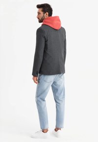 Grauer Blazer mit roter Kapuze, schlanker Schnitt, strukturierter Stoff und zwei Knöpfen. Kombiniert mit hellblauen Jeans und weißen Sneakern.