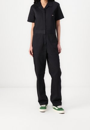 Personne portant une combinaison noire à manches courtes avec col, et des baskets vertes et blanches, debout devant un fond blanc uni.