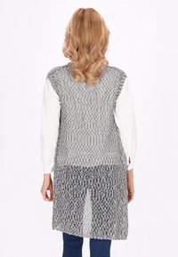 Cardigan lung gri, cu un design de tricot texturat, deschis în față și cu spatele mai lung, purtat peste un top alb cu mânecă lungă și pantaloni albaștri.