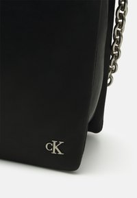 Borsa a tracolla nera in pelle con finitura testurizzata, tracolla in catena argentata e un piccolo logo argentato "cK" nell'angolo inferiore frontale.