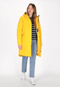 Gelbe wasserdichte Jacke mit Kapuze, Reißverschluss, zwei Fronttaschen und grauem Innenfutter. Kombiniert mit einem schwarz-weißen gestreiften Oberteil und Jeans.