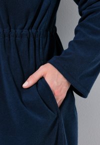 Šaty z námořnické modré tkaniny se skládající z elastického pasu, dlouhými rukávy a boční kapsou s hladkým povrchem. Ruka umístěná v kapse.